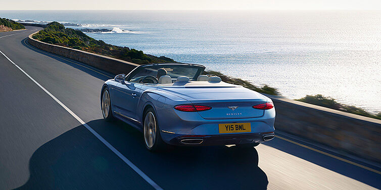 Bentley New Delhi New Bentley Continental GTC Azure convertible rear view in Light Windsor blue paint with 22 inch ten spoke wheels driving dynamically on an ocean road in the sunshine